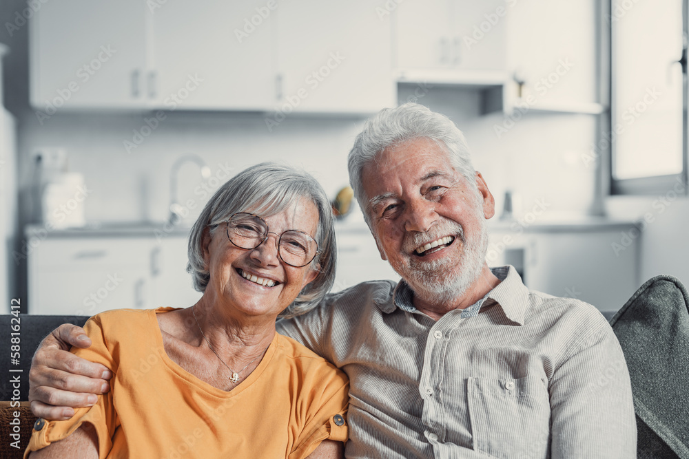 Headshot portrait of smiling elderly 60s husband and wife sit relax on couch hugging cuddling, happy mature old couple rest on sofa in living room embrace look at camera show love and care. Headshot portrait of smiling elderly 60s husband and wife sit relax on couch hugging cuddling, happy mature old couple rest on sofa in living room embrace look at camera show love and care.