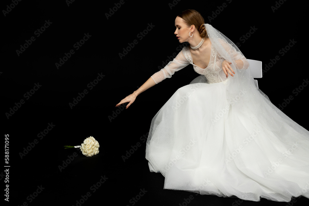 Naklejka premium A beautiful charming girl, the bride in a luxurious and elegant white wedding dress, reaching their hand to a graceful bouquet of white roses lying next to her. Isolated on a black background.