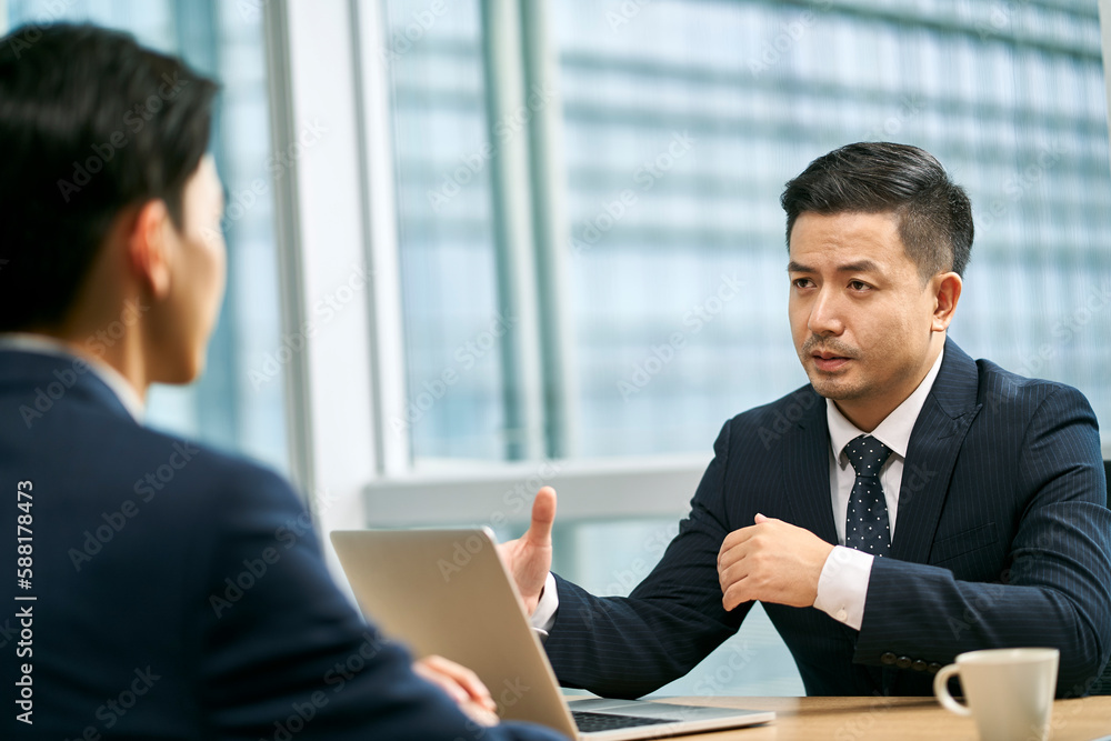 two asian corporate executives sitting at desk discussing business in office two asian corporate executives sitting at desk discussing business in office