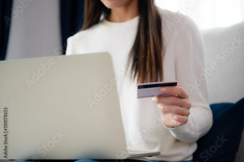 Young asian woman sitting on sofa in living room, makes online banking payments through the internet from bank card on computer laptop. Shopping online on notebook with credit card