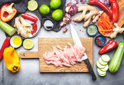 Food background. Sliced chicken fillet and chef knife on cutting board., vegetables, ginger, lime, chili pepper, garlic, soy sauce and rice vinegar on gray kitchen table. Asian stir fry ingredients