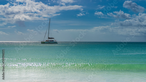 Tropical idyll. A white yacht is sailing on a calm turquoise ocean. A mast on a background of blue sky and clouds. The wave rolls towards the shore. Glare on the water. Seychelles.