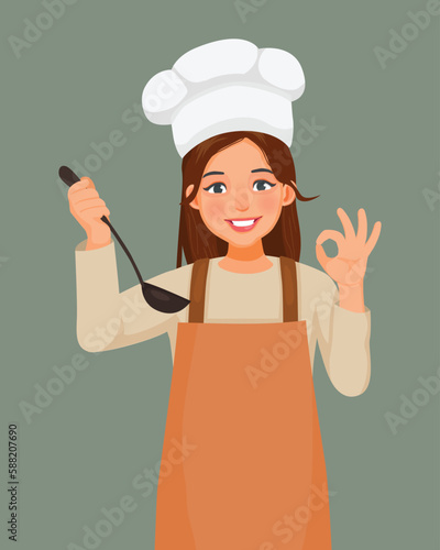 Young woman in chef hat and apron holding soup spoon showing okay sign
