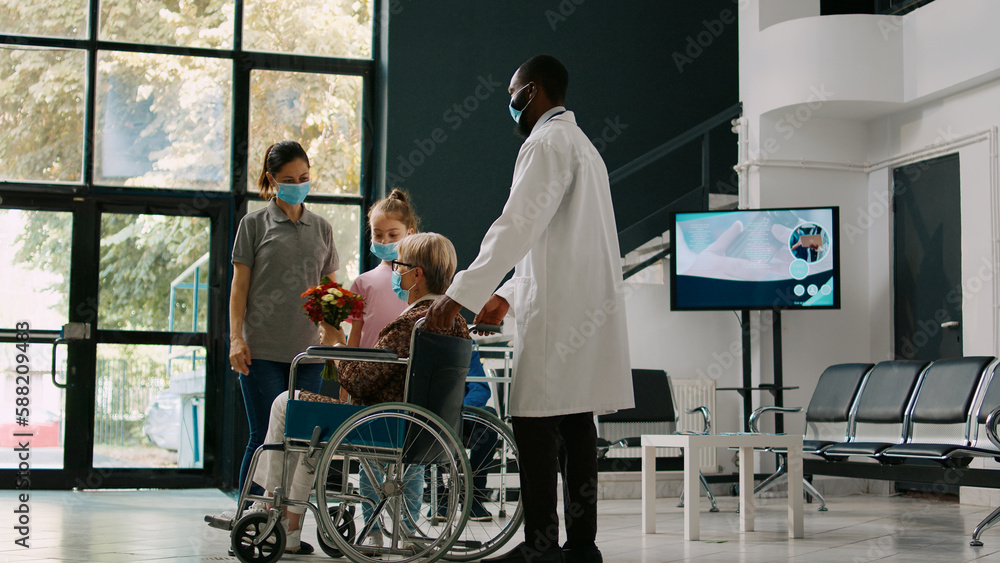 Little girl visiting senior woman in wheelchair during covid 19 pandemic, bringing flowers to elderly patient with disability. Mother and child coming to see person with physical impairment.