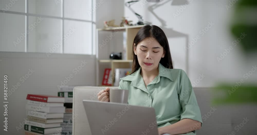 Handheld shot, Asian young woman sitting and drink beverage in mug, she smile with happy while use laptop computer in living room at home Handheld shot, Asian young woman sitting and drink beverage in mug, she smile with happy while use laptop computer in living room at home