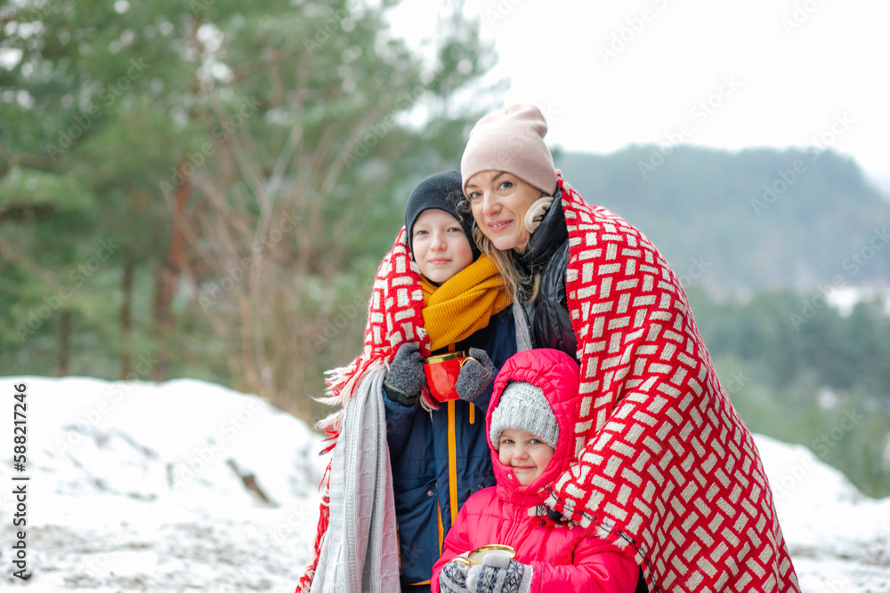 Cute, cozy family of woman, teen boy and little girl in warm clothes, hats, wrapped in blanket drink hot hot chocolate