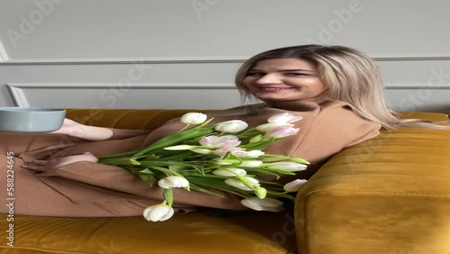 Happy pregnant woman drink coffee with bunch of fresh white tulips. Vertical female home video. Breakfasr on sofa. photo