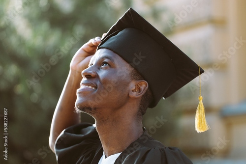 Portrait closeup of afro american student in graduation mantle and hat standing outdoors and with pleasure looking up. Job search, graduate from university, start in life, thoughts about future.