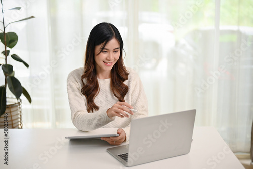 Beautiful young Asian woman using her tablet and laptop in her home office.