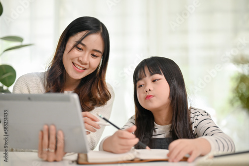 Kind and friendly Asian female sister teaching math to her little sister, looking at tablet screen