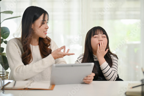 Adorable little Asian girl yawning while her mom is teaching math on tablet at home.