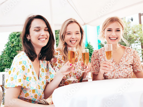 Three young beautiful smiling hipster female celebrating. Carefree women posing at summer terrace veranda cafe in the street.Positive models drinking champagne. Enjoying their vacation. Talking