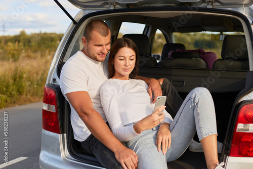 Photo of loving young family sitting in car trunk on road, man and woman wearing white shirts, making selfie, finding the shortest way while having break, using phone navigator. © sementsova321