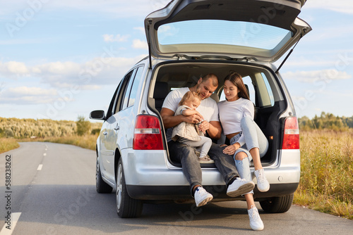 Portrait of happy family traveling together by car, resting, expressing positive emotions, sitting in automobile trunk with their infant daughter, enjoying moments.