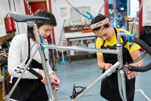 two girls checking the condition of a bicycle in a multi-repair workshop, multi-ethnic female mechanics repairing a sustainable vehicle, bike hung up for supervision