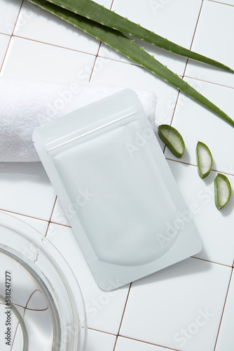 A facial sheet mask packaging mockup, white towel and fresh aloe vera leaves on white tile floor. Background for advertising cosmetic of aloe vera extract with bathroom concept.