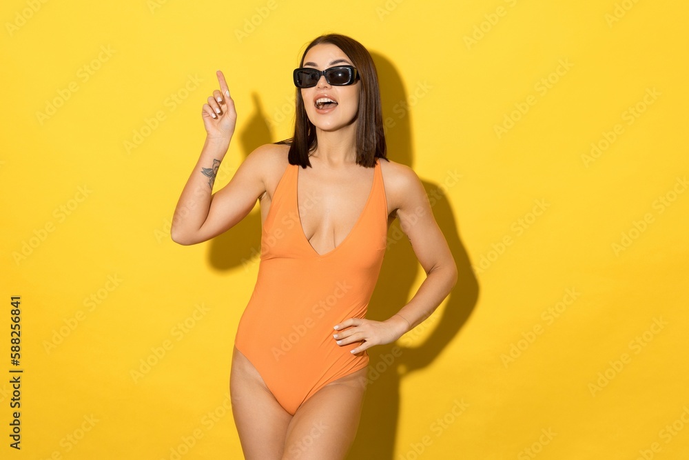Sexy young woman in an orange swimsuit and sunglasses on a yellow background. The model points her finger up. Mockup. Advertisement
