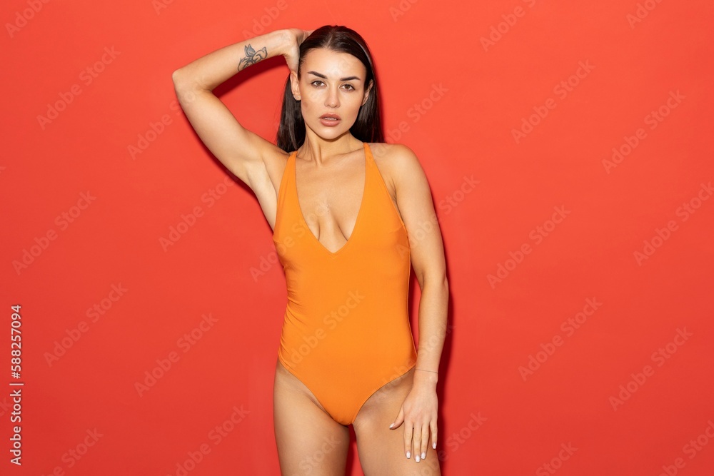 A tanned woman in an orange swimsuit on a red background. Attractive athletic figure. Summer vacation on the beach by the sea A tanned woman in an orange swimsuit on a red background. Attractive athletic figure. Summer vacation on the beach by the sea