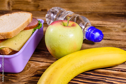 Ripe apple, banana, bottle of water and lunch box with sandwiches on wooden table