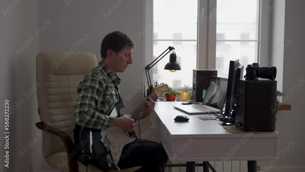 Young man playing electric guitar sitting by the desk in a room Young man playing electric guitar sitting by the desk in a room