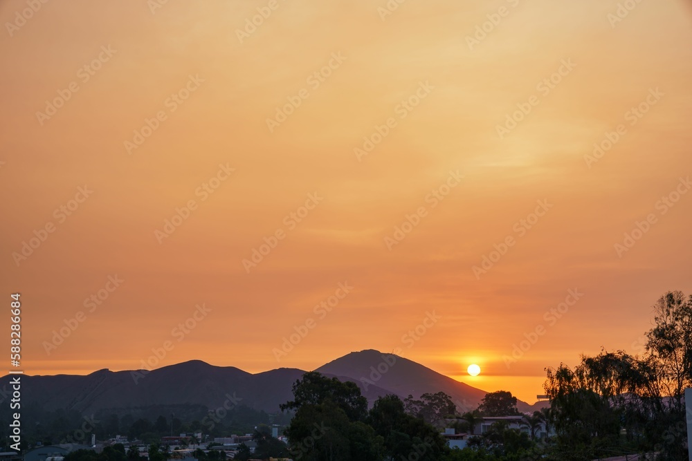 Small bright sun in light orange sunset sky shining behind mountains above houses and green trees