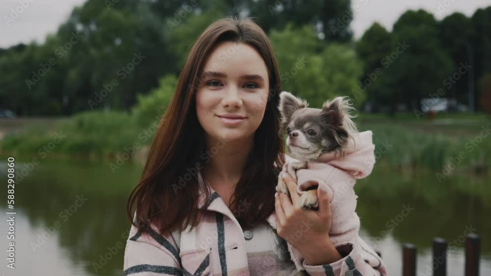 Portrait of the Young Beautiful Happy Woman, Carrying Her Lovely Small Chihuahua. Cute Girl Looking at the Camera on the Local Lake and Park Background. People and Dogs Friendship Concept Portrait of the Young Beautiful Happy Woman, Carrying Her Lovely Small Chihuahua. Cute Girl Looking at the Camera on the Local Lake and Park Background. People and Dogs Friendship Concept