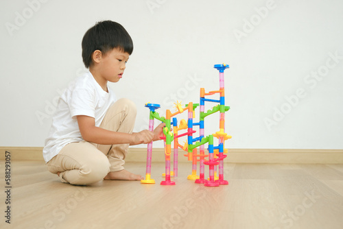 Cute Asian kindergarten boy child sitting on floor having fun playing with colorful plastic Marble Run Track Toy Set, Activities at home, Educational and Fun toys for young kid, isolated on white wall