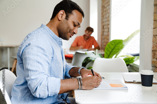 Smiling indian man writing down notes while sitting in coworking