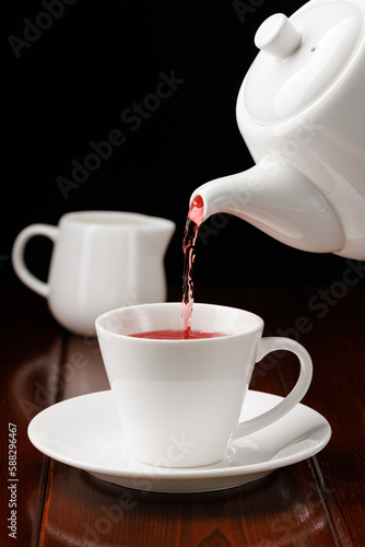 Red tea is poured from a white teapot into a white cup on a black background. Porcelain teaware. Side view. Selective focus.
