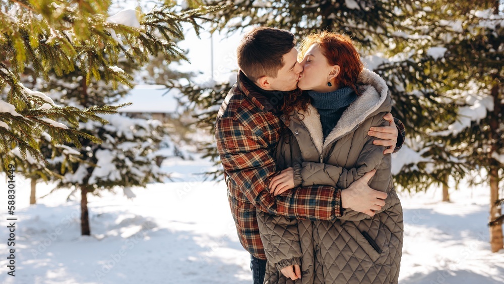 A view of a kissing man and woman standing in the midst of snow-covered trees. A young man hugs and kisses his lady during a date in a snowy park. A view of a kissing man and woman standing in the midst of snow-covered trees. A young man hugs and kisses his lady during a date in a snowy park.