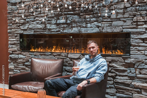 Fototapeta Naklejka Na Ścianę i Meble -  Portrait of man with wineglass sitting in armchair in front of illuminated fireplace of luxury hotel during vacation, luxury travel concept