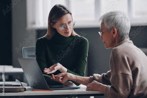 Confident financial advisor pointing laptop while discussing options with senior woman in office