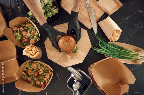Close up view of burger, French fries and vegetables. Man is packing food into the paper eco boxes. Indoors, restaurant
