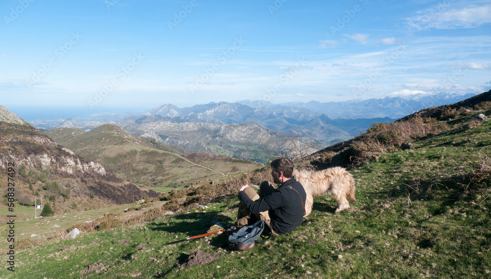 Hombre joven barbudo y perro sentado  en ladera de montaña de Asturias