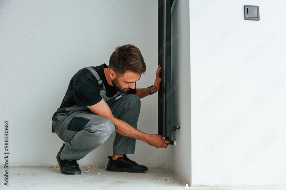 Plumber worker installing heating radiator in empty room of a newly built house