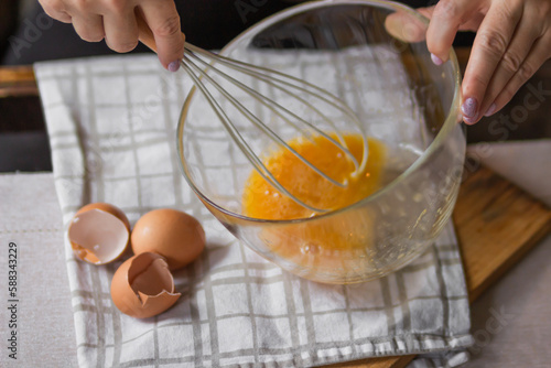Elderly woman beats eggs with a wisk in a glass bowl. Prepare for making christmas pie or pancakes on kitchen table.