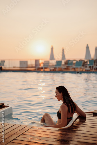 A girl in a black swimsuit sits at the entrance to an outdoor pool without people. The sun is setting. In the evening at sunset. Looks into the distance © Ilya.K