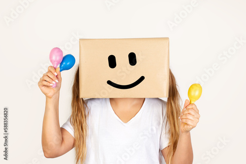 Mujer sonriente con una caja de cartón en la cabeza sosteniendo unas maracas de colores sobre un fondo blanco liso y aislado. Vista de frente y de cerca. Copy space 