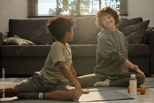 Young smiling woman in activewear looking at her son during exercise on mat while both sitting on the floor of living room by grey couch