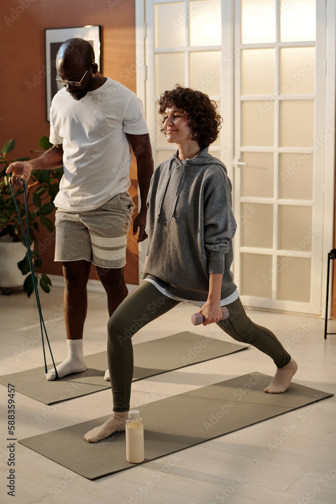 Young woman with dumbbell in hand doing stretching exercise for legs while standing on mat against her husband with resistance band Young woman with dumbbell in hand doing stretching exercise for legs while standing on mat against her husband with resistance band