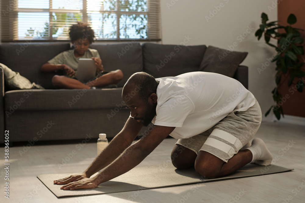 Side view of young African American man stretching arms forwards while standing on knees on the floor of living room during workout Side view of young African American man stretching arms forwards while standing on knees on the floor of living room during workout