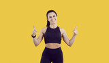 Portrait of smiling toned female athlete in sportswear isolated on yellow studio background show thumbs up. Active overjoyed young woman coach or trainer recommend sport and physical activity.