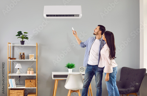 Young family couple using their modern air conditioning system at home. Happy husband and wife setting up the temperature on their white AC on the wall in the living room. Air conditioner concept photo