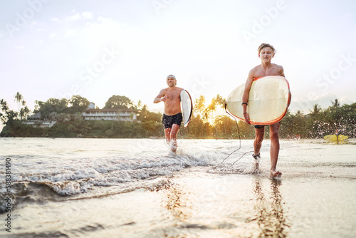 Photography Young teen boy with a surfboard walking with his father by ocean sandy beach after surfing with beautiful sunset background