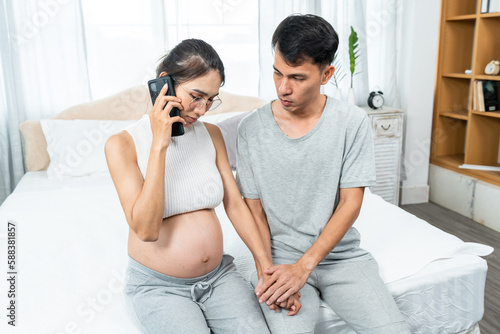 Asian pregnant woman talking on mobie phone with the doctor and having stress. husband held hand and gently stroked his wife's hand, relieving stress and consoling his wife to encourage each other.
