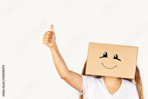 Mujer con caja de cartón en la cabeza con gesto positivo y feliz y el dedo pulgar de la mano hacia arriba OK sobre fondo blanco liso y aislado. Vista de frente y de cerca. Copy space photo