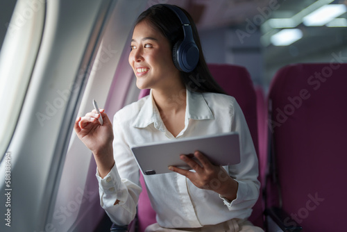 Young Asian attractive business woman passenger sitting on business class luxury plane while wearing wireless headphone for relax and working with tablet during the flight
