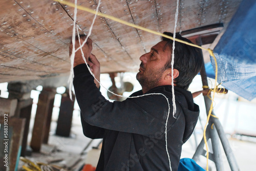 A middle-aged or young man working on the hull of a boat - putting cotton caulk into the cracks between wooden planks (caulking) photo
