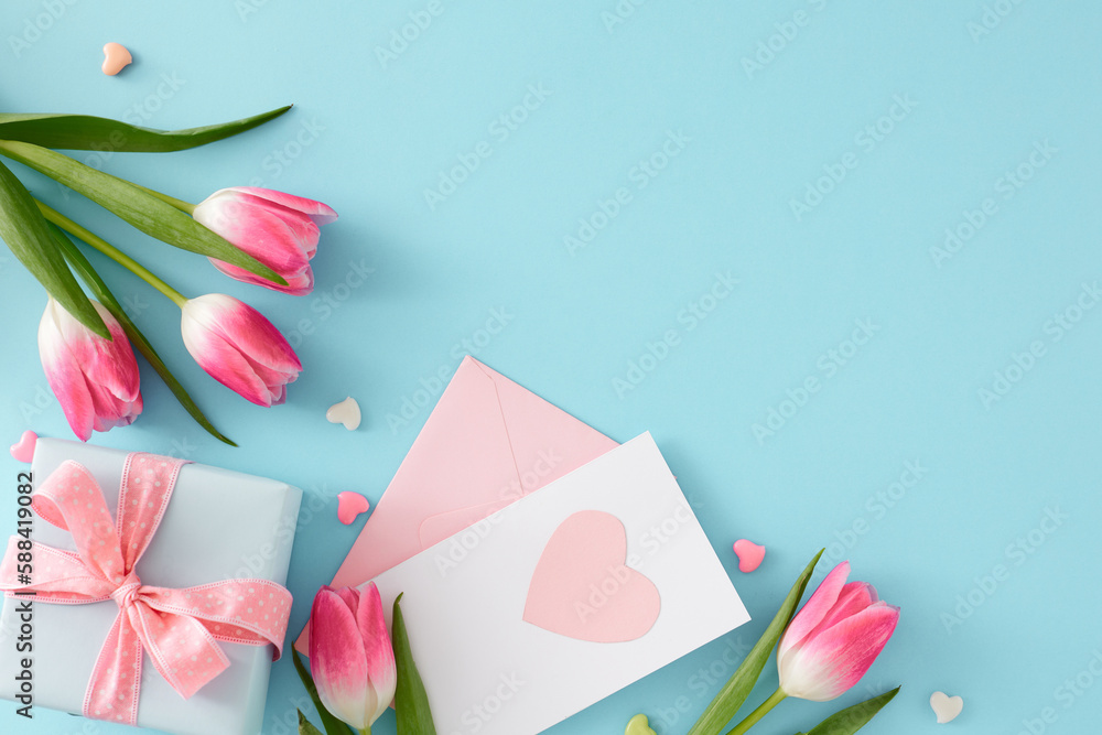 Women's Day atmosphere concept. Flat lay photo of gift box with bow postcard hearts baubles pink tulips flowers on isolated pastel blue background with copyspace Women's Day atmosphere concept. Flat lay photo of gift box with bow postcard hearts baubles pink tulips flowers on isolated pastel blue background with copyspace
