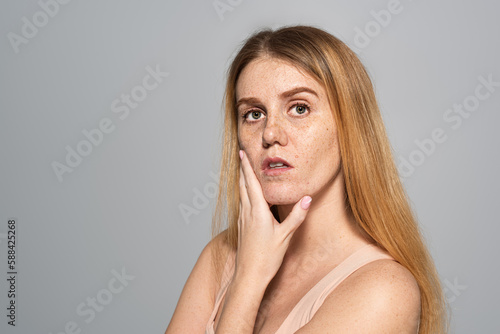 Young woman with freckles on face touching skin isolated on grey.